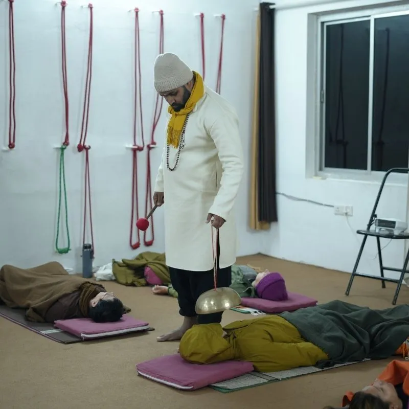 A practitioner performing a Himalayan singing bowl sound healing session on participants lying in a meditation studio. The healer holds a brass singing bowl over a student during a sound bath therapy class.