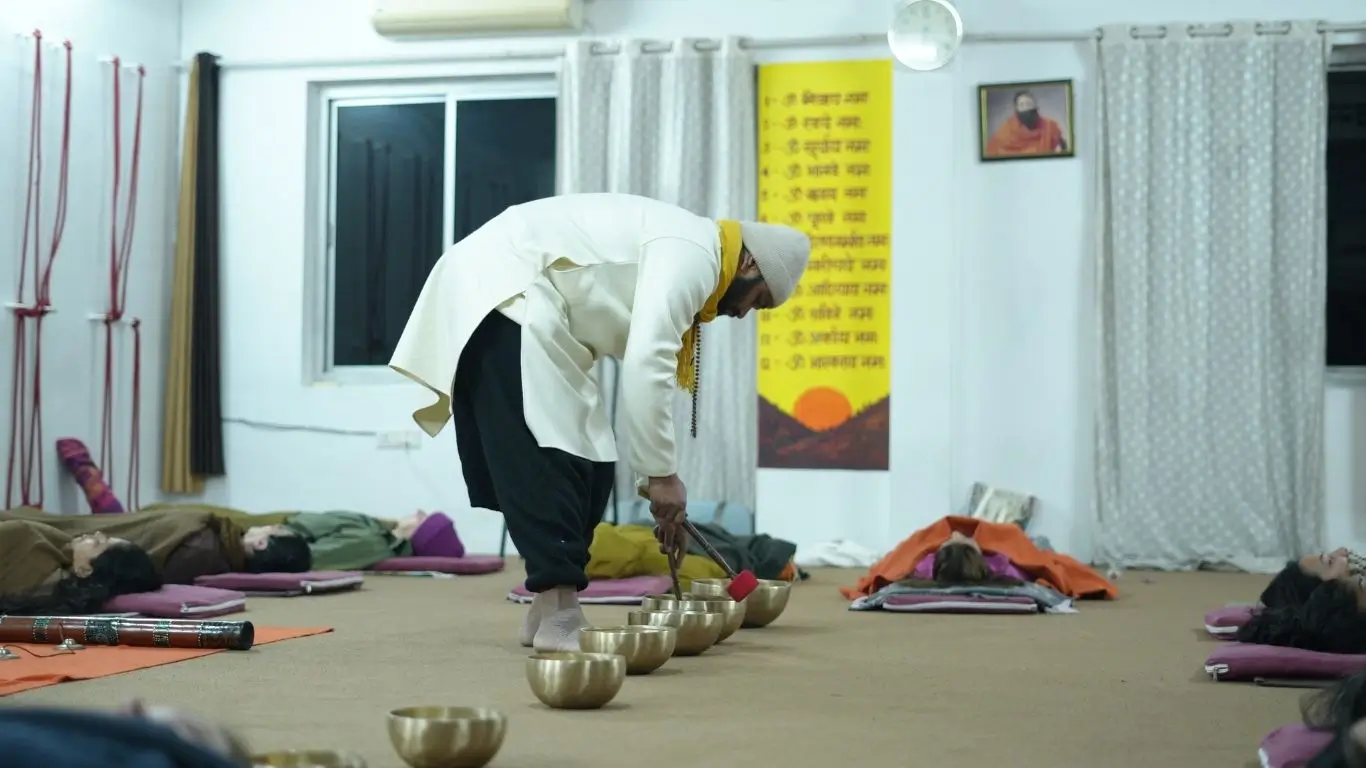 Sound healing teacher playing Tibetan singing bowls during a sound bath session in Rishikesh, India, with students relaxing on yoga mats.
