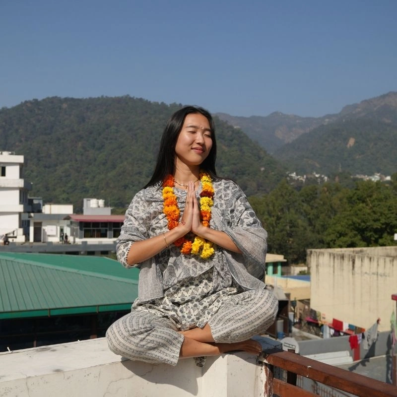 A female student practicing meditation in a seated prayer pose during a 100 hour Kundalini yoga teacher training in Rishikesh, India, with the Himalayan foothills in the background.