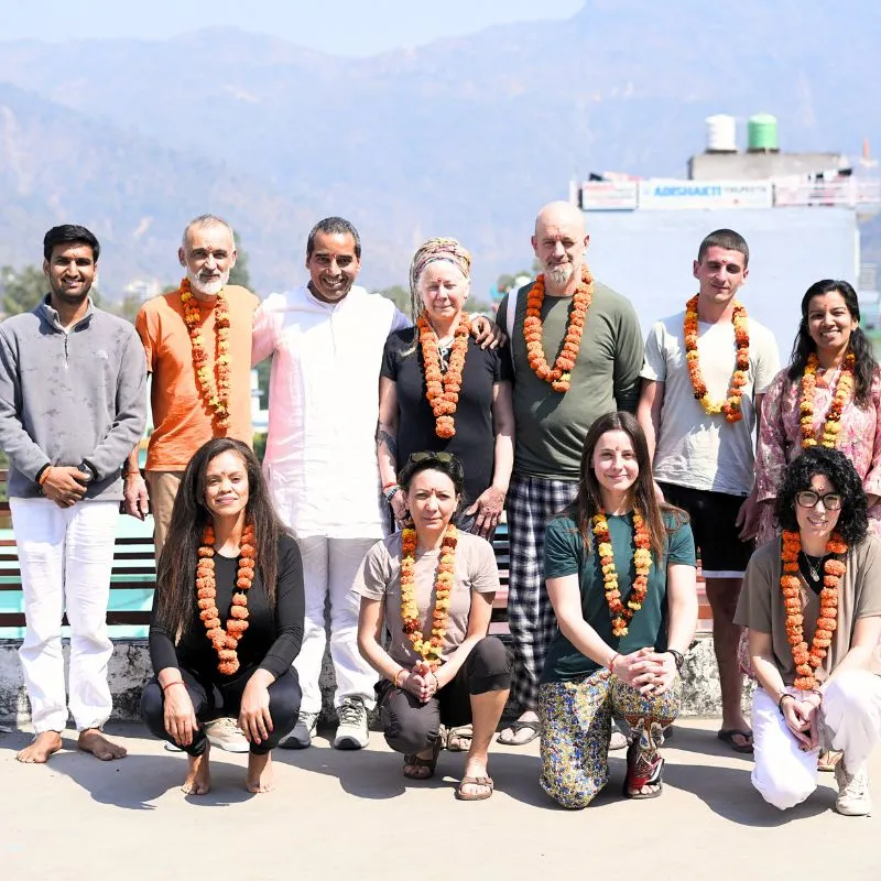 International students and teachers celebrating completion of 100 hour yoga teacher training in Rishikesh India with traditional flower garlands and Himalayan mountain background.