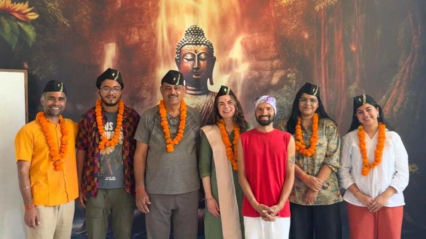 A diverse group of smiling students and teachers wearing traditional marigold garlands and black caps, celebrating the completion of a 200 hour Kundalini yoga teacher training in Rishikesh, India, standing in front of a Buddha backdrop.