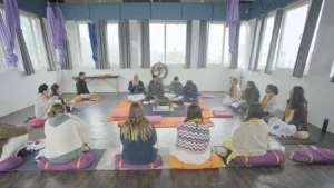 A group of students participating in a traditional opening ceremony or chanting session during a yoga teacher training for beginners in a bright Rishikesh studio.