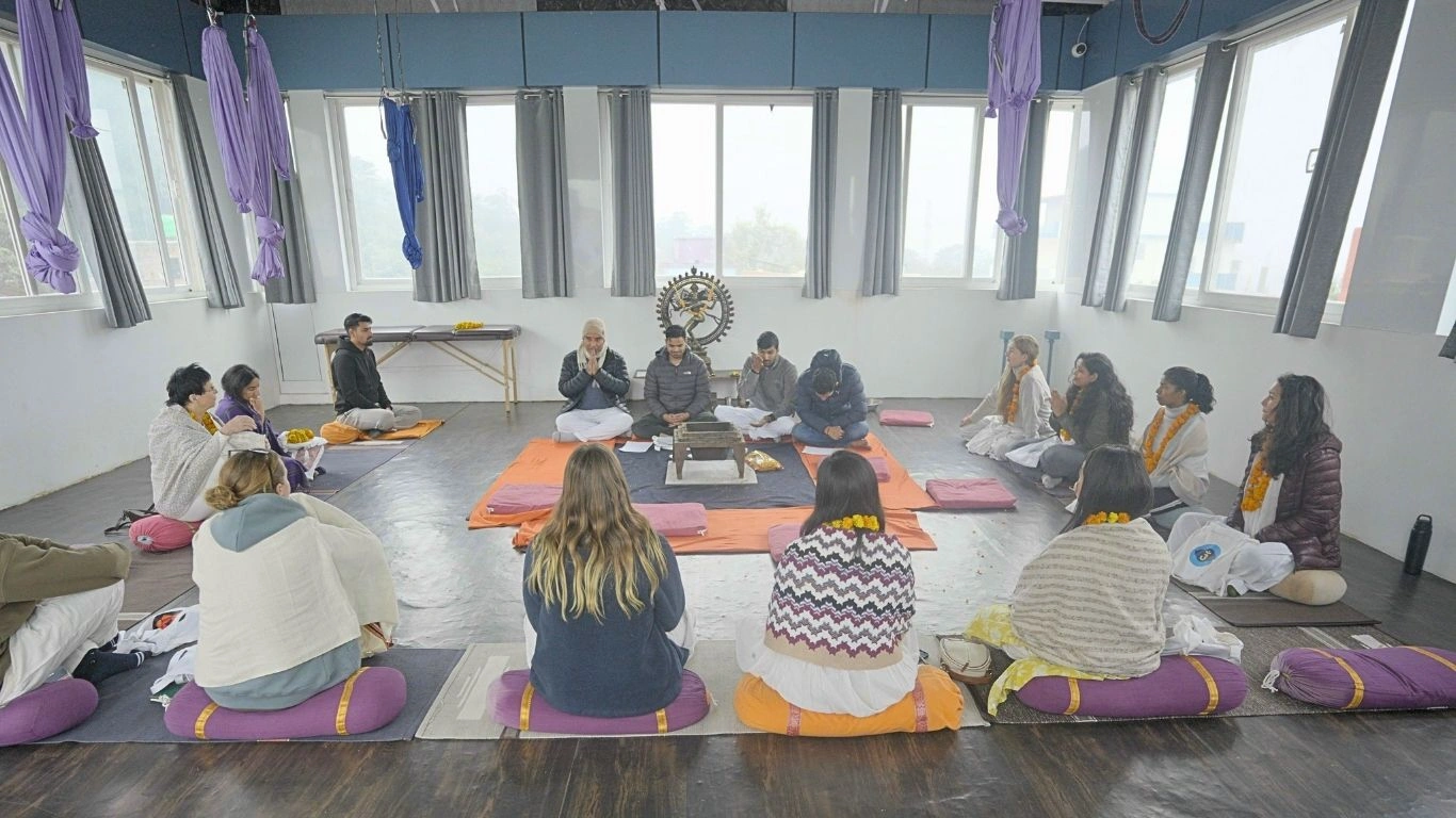 A group of students participating in a traditional opening ceremony or chanting session during a yoga teacher training for beginners in a bright Rishikesh studio.