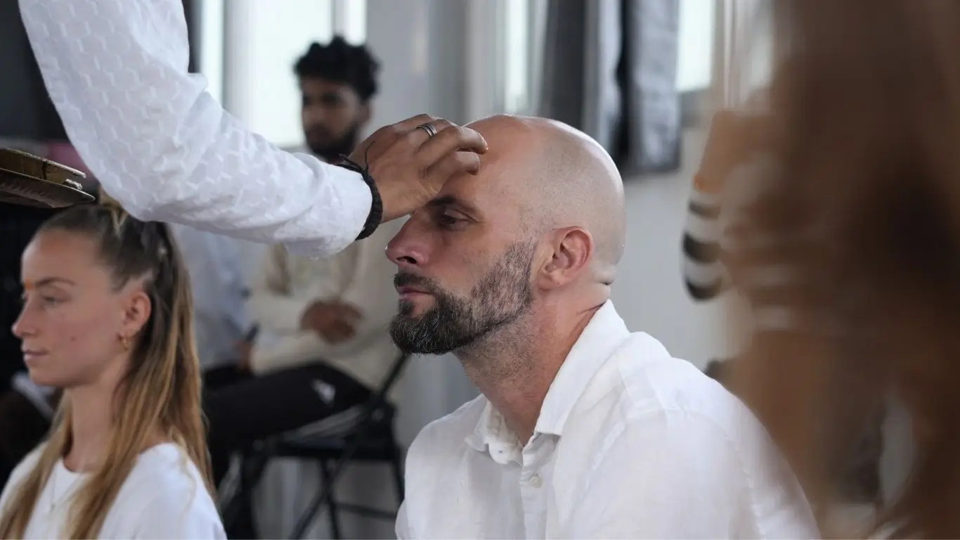 A close-up shot of a practitioner receiving a blessing or Tilak on the forehead during a ceremony at Rishikesh Yoga Valley. This ritual is a significant aspect of a traditional yogic lifestyle, emphasizing the opening of the third eye and spiritual focus. The practitioner is dressed in white, surrounded by others in a serene, meditative environment.