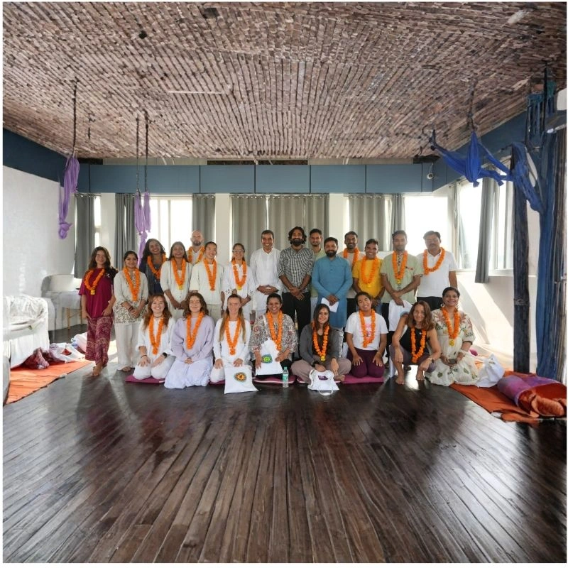A large group of yoga students and teachers posing together in a wooden-floored studio with aerial yoga silks during a 100 hour Kundalini yoga teacher training in Rishikesh, India.