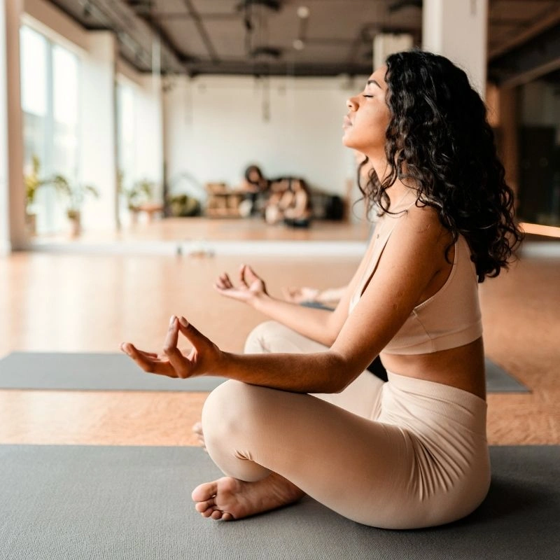 Side view of a woman with curly hair practicing meditation in a yoga studio during a 200 hour Kundalini yoga teacher training in Rishikesh, India, with other students in the background.