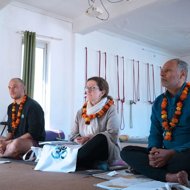 Three diverse students wearing traditional orange garlands and seated in a yoga shala during a 100 hour Kundalini yoga teacher training in Rishikesh, India.
