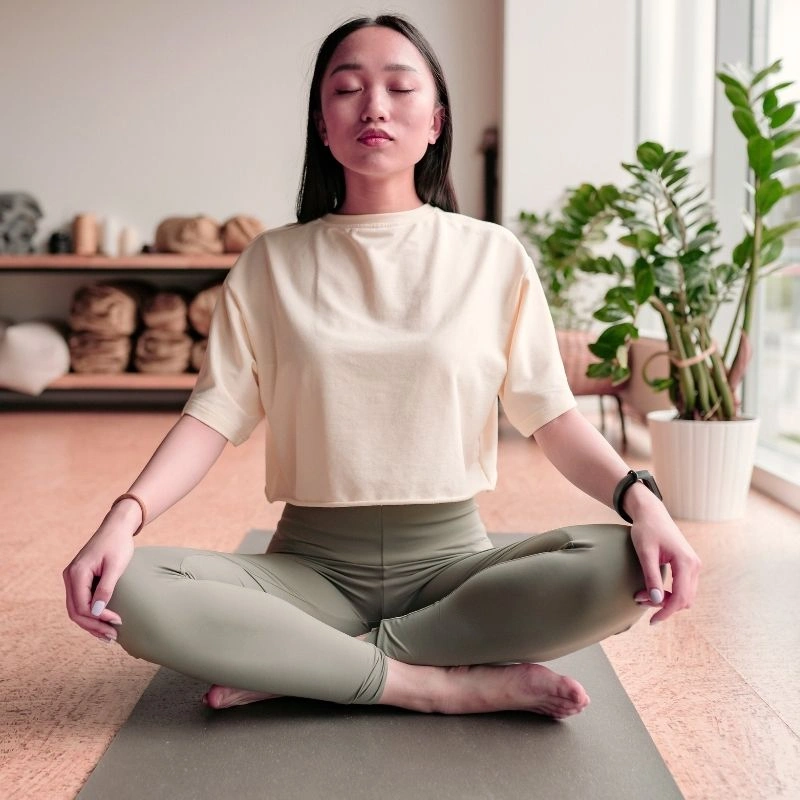 A young woman sitting in a cross-legged meditative pose on a yoga mat during a 200 hour Kundalini yoga teacher training in Rishikesh, India, within a bright and modern yoga studio.A young woman sitting in a cross-legged meditative pose on a yoga mat during a 200 hour Kundalini yoga teacher training in Rishikesh, India, within a bright and modern yoga studio.