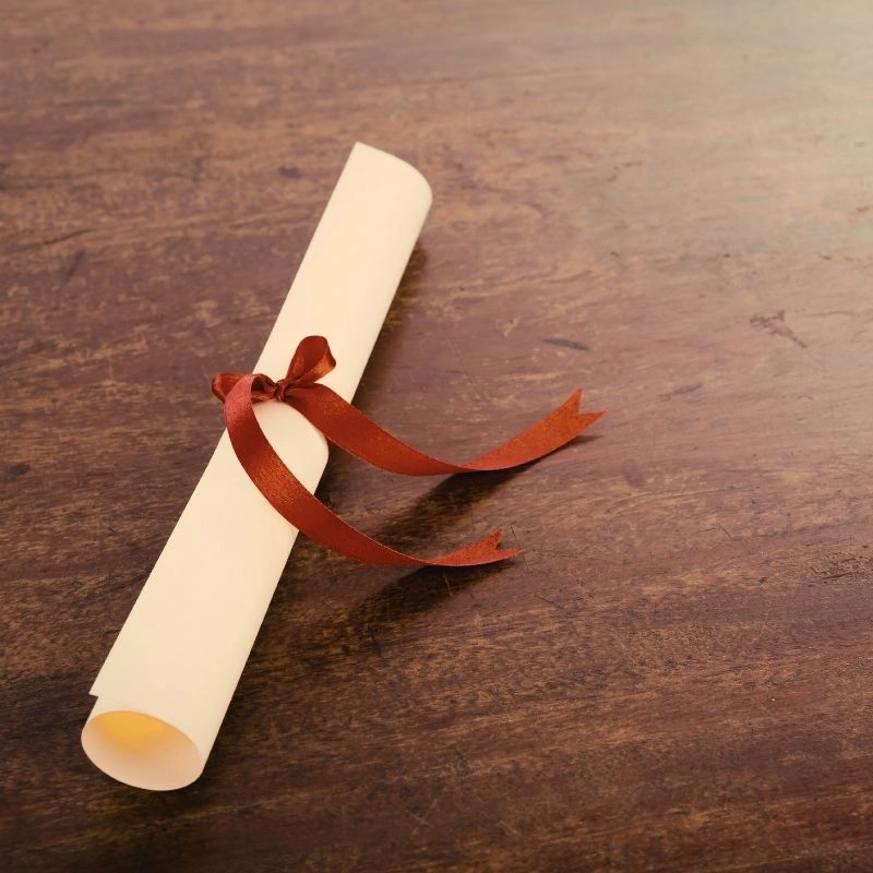 A rolled-up diploma tied with a red ribbon on a wooden table, representing the graduation certificate from a 200 hour Kundalini yoga teacher training in Rishikesh, India.