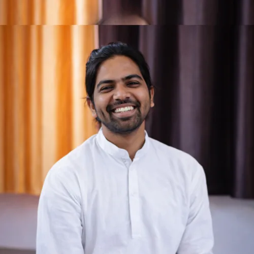 A smiling young male yoga instructor with long hair tied back, wearing a white traditional kurta, posing during a 50 hour yoga teacher training in Rishikesh India.