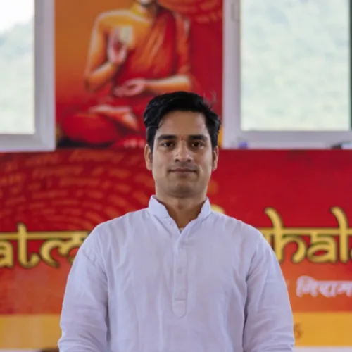 A portrait of an Indian male yoga teacher in a white traditional kurta standing in front of an orange spiritual banner during a 50 hour yoga teacher training in Rishikesh India.