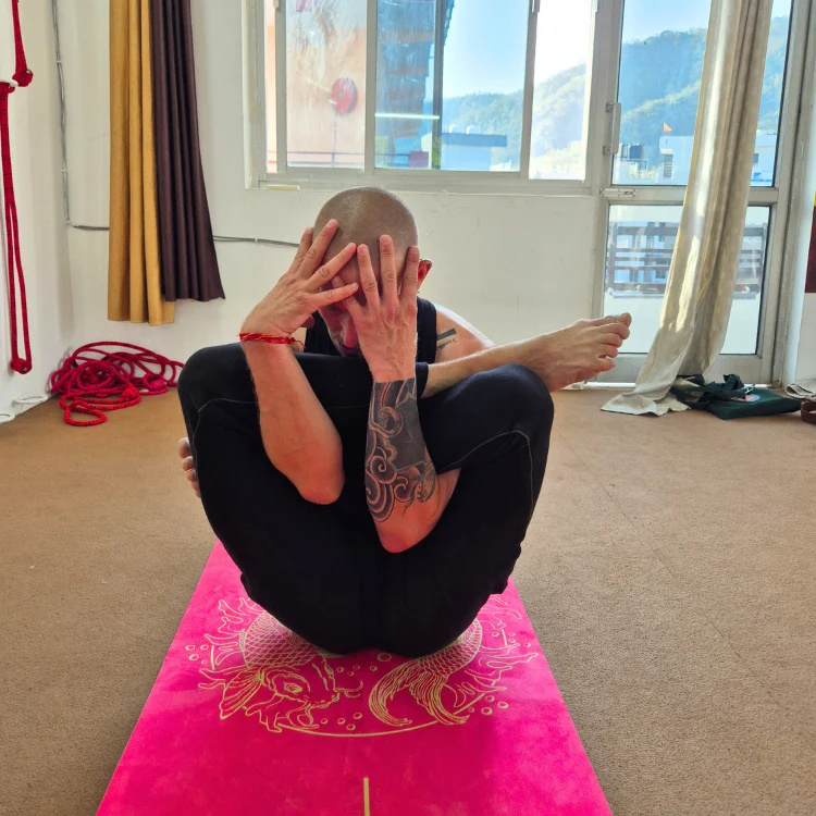 An advanced practitioner with arm tattoos demonstrating a deep leg-behind-head variation (Yoganidrasana or Garbha Pindasana variant) on a pink yoga mat during a 300 hour yoga teacher training in rishikesh india.