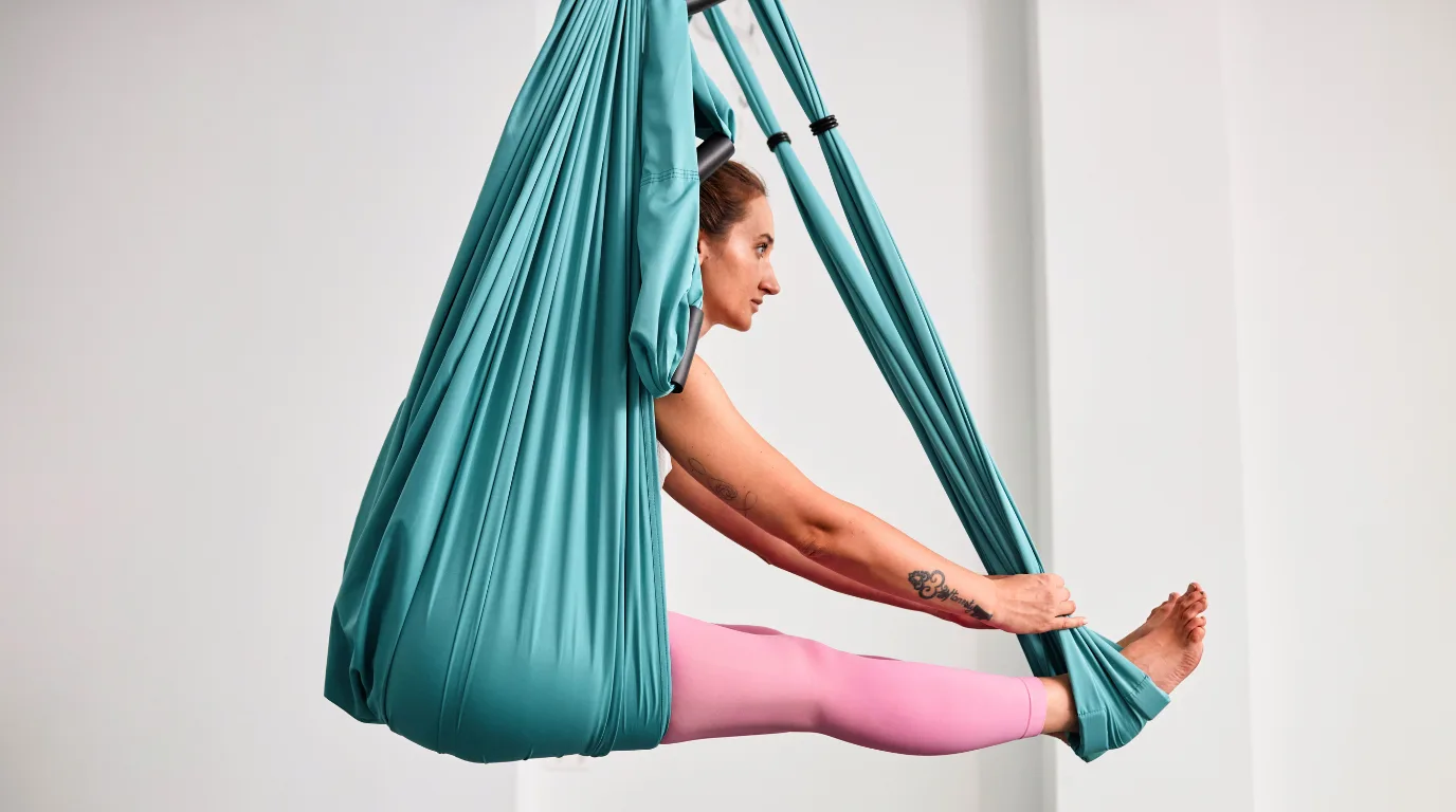 Woman practicing a seated stretch using a teal hammock during aerial yoga teacher training in Rishikesh, India.