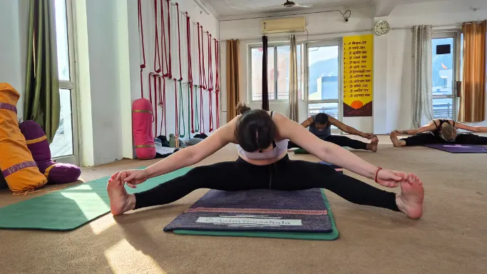 Students practicing a seated wide-angle forward fold pose (Upavistha Konasana) on yoga mats during an ashtanga session during yoga teacher training in rishikesh.
