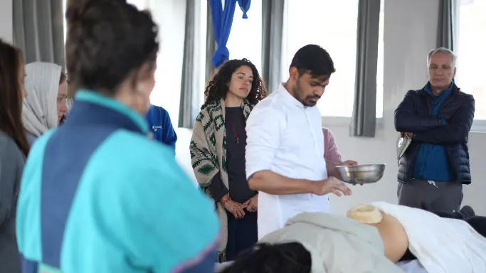 An instructor demonstrating a traditional oil treatment (Kati Basti) on a patient's lower back while a group of students watches intently during an ayurvedic massage during 10 days ayurveda & yoga retreat in rishikesh.