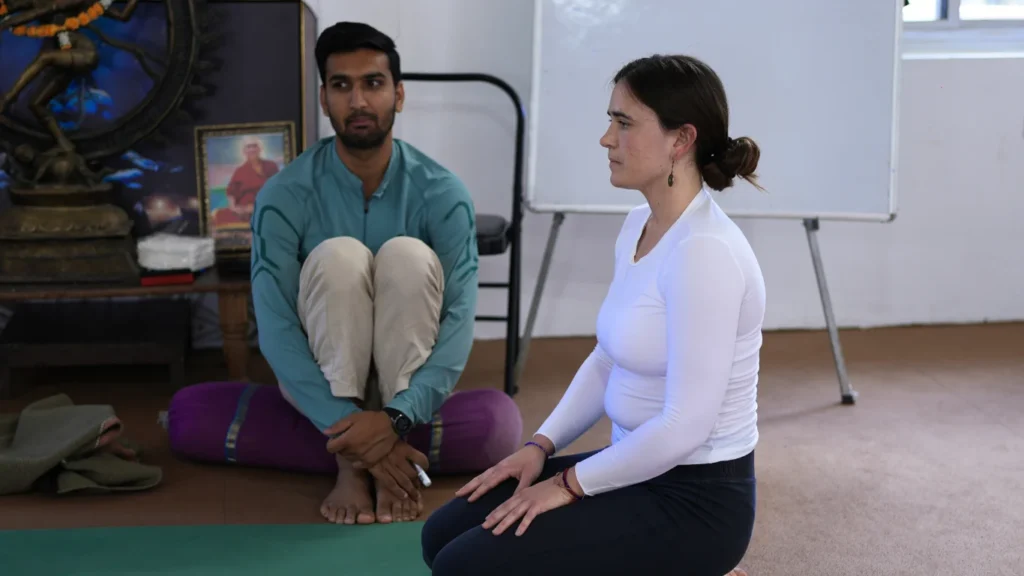 A female student practicing a seated meditation pose while a male yoga instructor observes closely, illustrating the answer to: can beginners join a 200 hour yoga teacher training