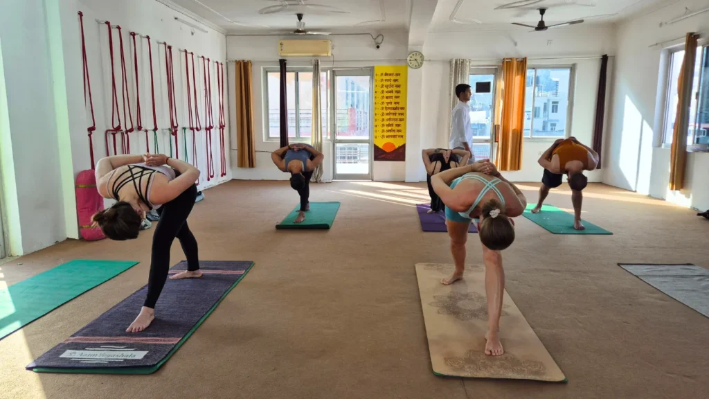 A group of yoga students practicing Pyramid Pose (Parsvottanasana) in a bright studio space under an instructor's guidance, answering: can beginners join a 200 hour yoga teacher training