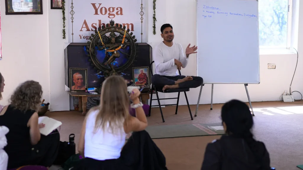 An Indian yoga instructor teaching a theory class on a whiteboard at a yoga ashram while students take notes, demonstrating if can beginners join a 200 hour yoga teacher training course.