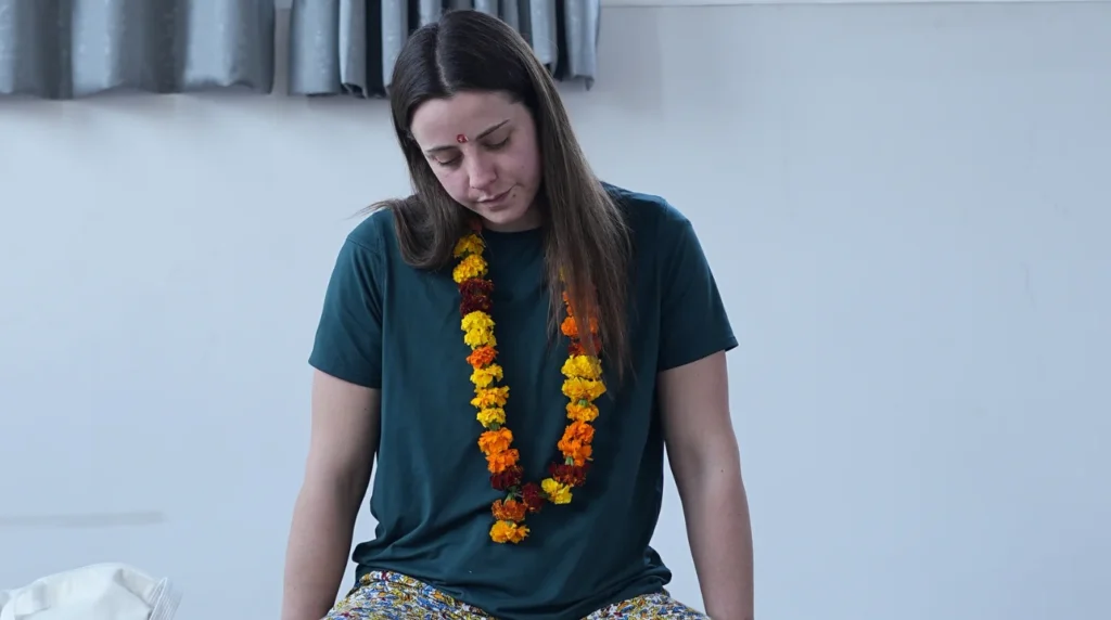 Young woman wearing a traditional marigold garland sitting in quiet meditation with her eyes closed during a 200 hour yoga teacher training session