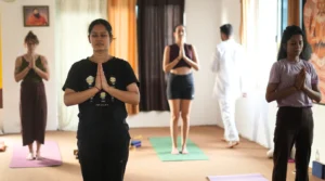 A group of diverse students standing in Tadasana with eyes closed and hands in Anjali Mudra prayer position, demonstrating dedicated mindfulness during a 200-hour yoga teacher training class.