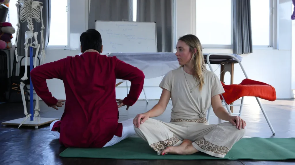 A yoga student sitting cross-legged on a mat, observing an instructor demonstrate posture alignment in a classroom equipped with an anatomical skeleton model during a 200-hour yoga teacher training.