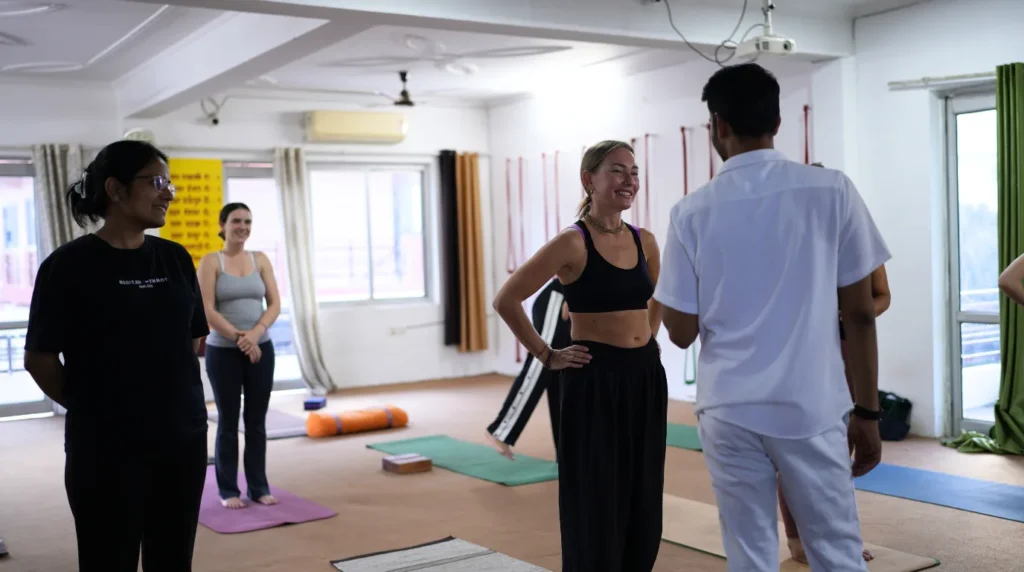 A smiling female yoga student interacting with a teacher and classmates during a group discussion and practice session in a 200-hour yoga teacher training classroom.