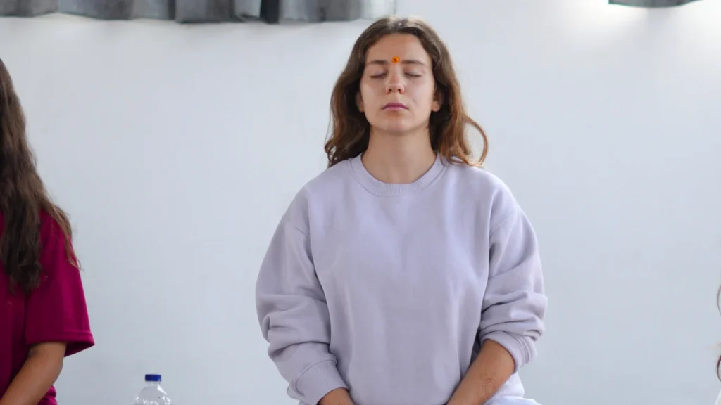 A young woman with a bindi meditating with her eyes closed in a white sweatshirt, showcasing how difficult is 200 hour yoga teacher training to master internal stillness.