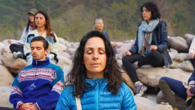 A group of people sitting cross-legged with eyes closed on rocks by a riverbank outdoors, practicing mindfulness during a meditation session during 5 days yoga & meditation retreat in rishikesh.