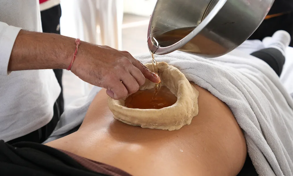 A therapist pouring warm herbal oil into a herbal dough ring on a person's lower back for a Kati Basti treatment during an Ayurveda module of a yoga course in Rishikesh.