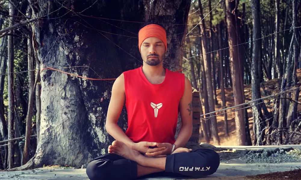 Yoga instructor Kamal Negi wearing an orange headband and red tank top, sitting in a cross-legged meditative lotus pose during an outdoor yoga course in Rishikesh surrounded by trees.