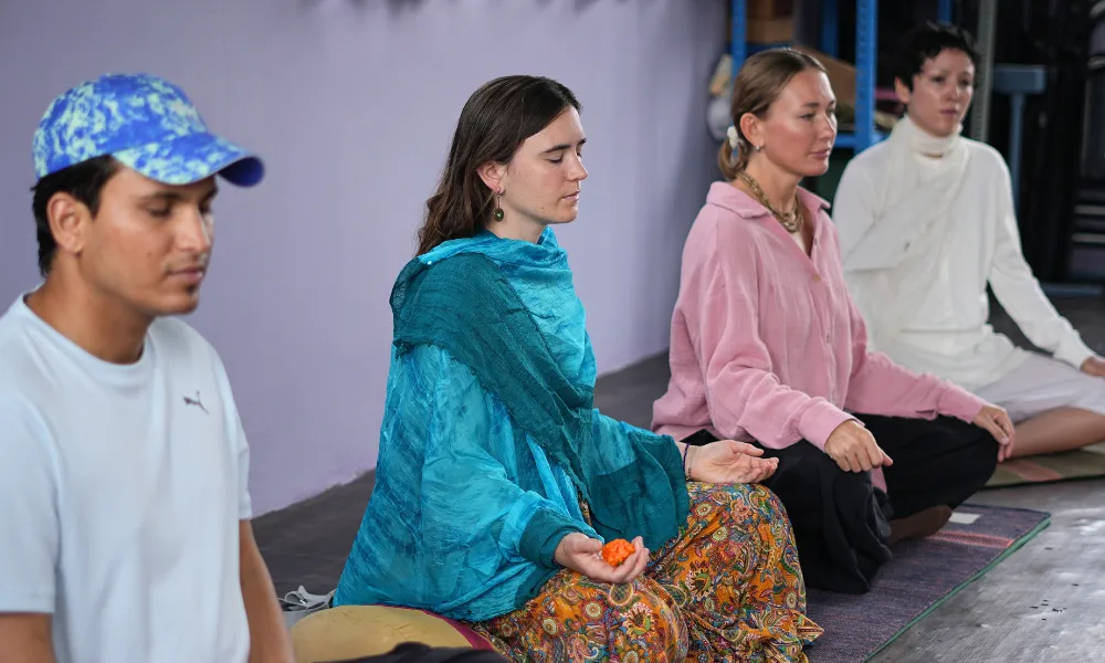 A row of male and female students sitting in a cross-legged meditative pose with their eyes closed during a yoga course in Rishikesh inside a calm studio room.