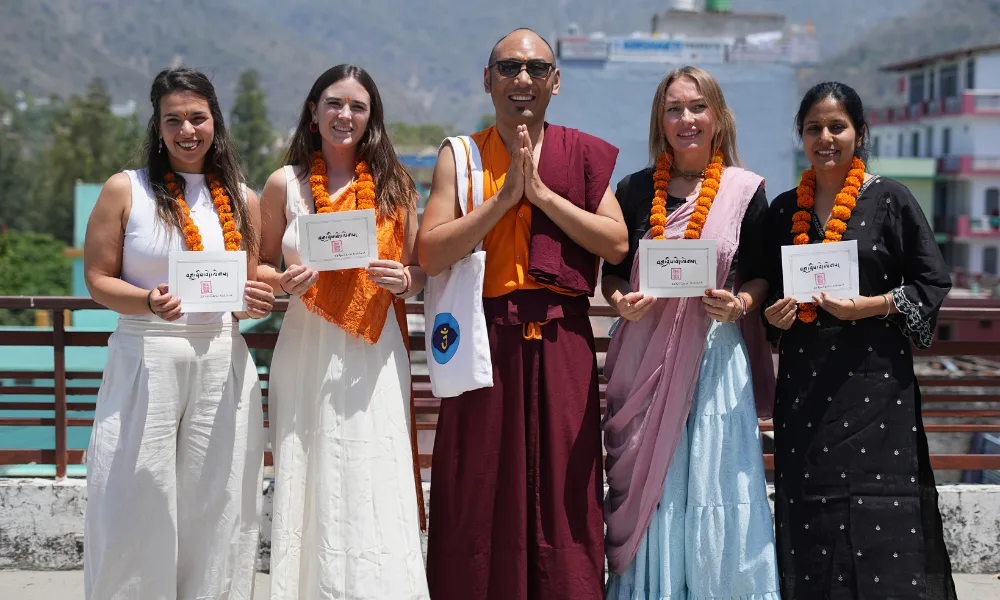 A group of international students holding graduation cards alongside their teacher in traditional robes, wearing marigold garlands on a outdoor rooftop during a yoga course in Rishikesh.