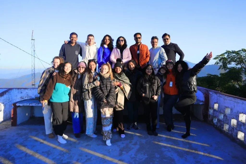 A diverse group of international yoga training students smiling together during an outdoor mountain excursion in India.