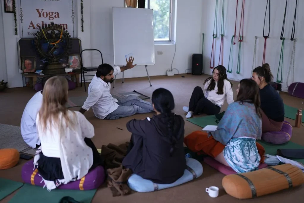 Yoga students sitting in a circle on cushions listening to a lecture on traditional vedic philosophy inside a yoga shala.