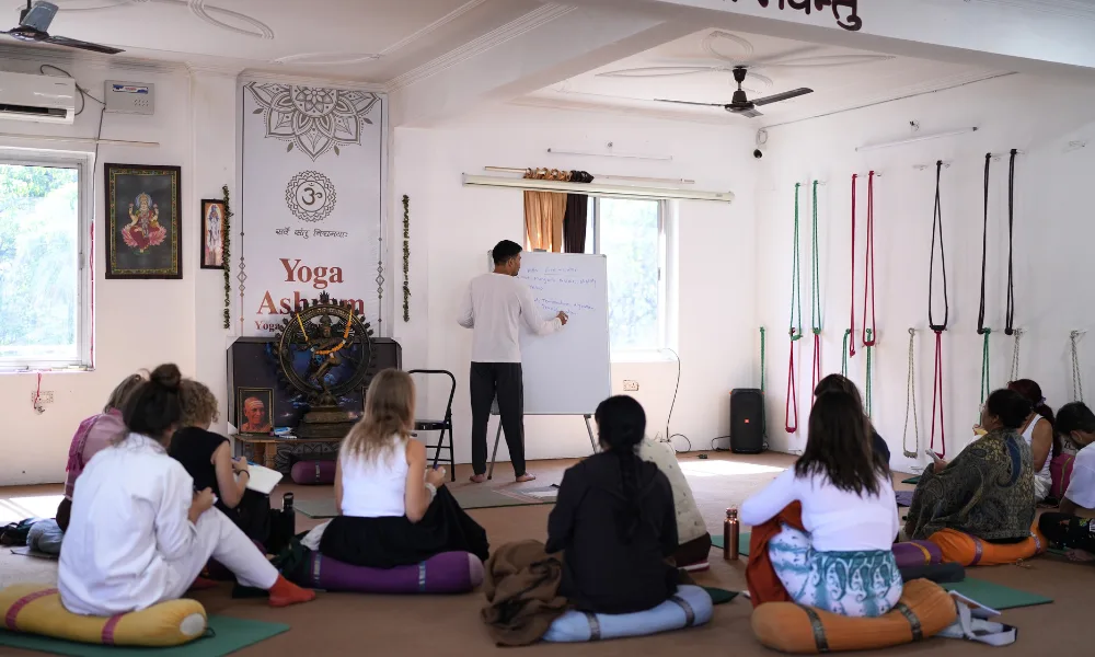 Students sitting on bolsters and taking notes during a philosophy lecture by an instructor writing on a whiteboard inside an authentic yoga school in Rishikesh.