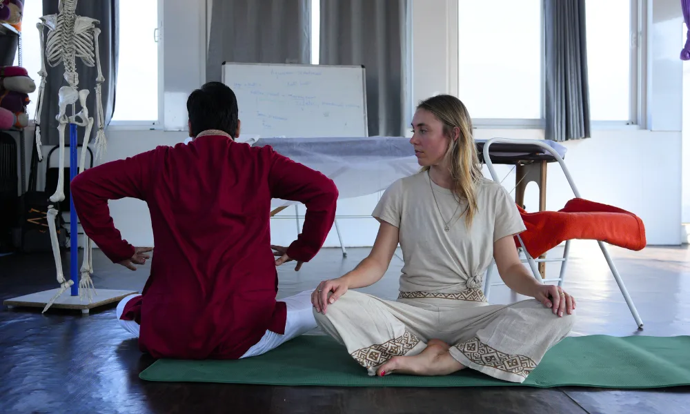An instructor in a red tunic and a female student sitting on a yoga mat during a yoga teacher training in Rishikesh anatomy class, with a human skeleton model and a whiteboard in the background.