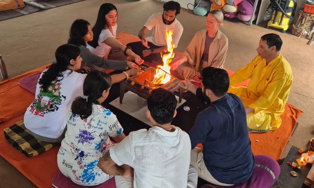 Students and a priest participating in a traditional sacred fire ceremony (Havan) during a yoga teacher training in Rishikesh, sitting in a circle around a burning fire pit.