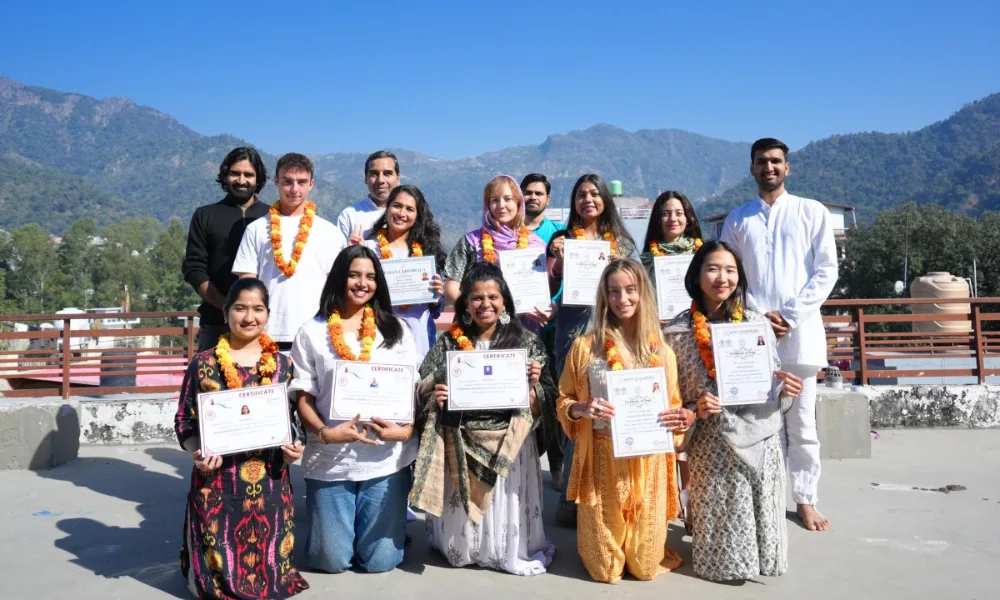 A group of international students proudly holding their graduation certificates after completing a yoga teacher training in Rishikesh, standing outdoors on a rooftop with the Himalayan mountains in the background.