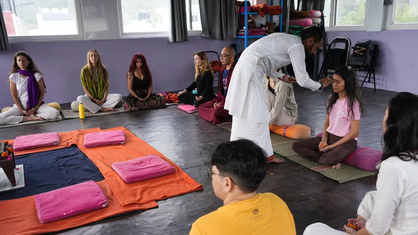 An instructor applying a traditional tilak blessing to a student's forehead while others sit in meditation cushions during a yoga teacher training in rishikesh opening ceremony.