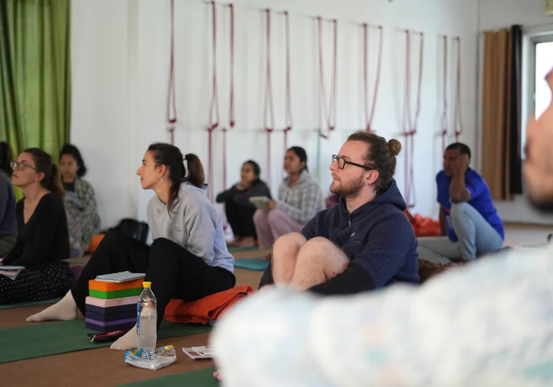 Students sitting on yoga mats with notebooks and props listening attentively during a yoga teacher training in rishikesh philosophy session.