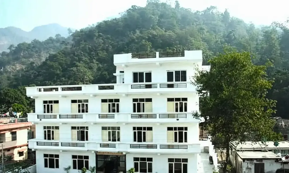 Exterior view of a multi-story white building hosting a yoga teacher training in Rishikesh, surrounded by lush green hills and mountains