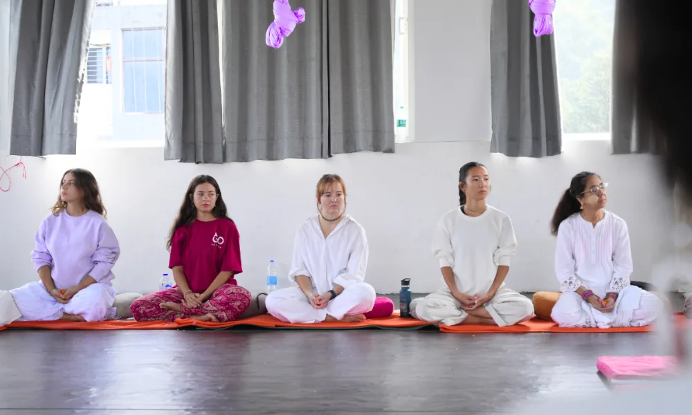 Students sitting in a cross-legged meditative pose during a yoga teacher training in Rishikesh, India, inside a bright studio with large windows