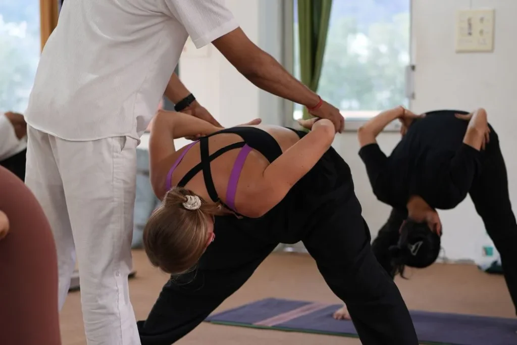 A senior yoga teacher provides hands on alignment correction to a student practicing a forward bend asana.