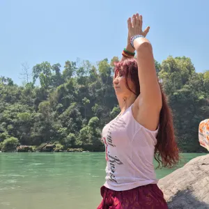 Woman practicing outdoor meditation with raised hands in a prayer pose by the sacred Ganges River during a 100 hour Kundalini yoga teacher training in Rishikesh.
