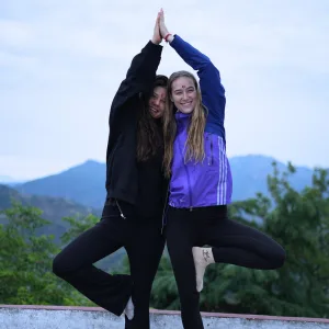 Two smiling female yoga students performing a partner tree pose against a scenic mountain backdrop during the 200 hour Kundalini yoga teacher training in Rishikesh.
