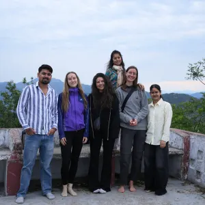 A group of yoga students and teachers posing together outdoors on a rooftop viewpoint with Himalayan mountains in the background during the 200 hour Kundalini yoga teacher training in Rishikesh.