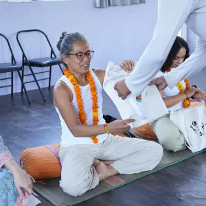 A smiling student wearing a orange marigold garland receiving a welcome kit bag during the opening ceremony of the 200 hour Kundalini yoga teacher training in Rishikesh.