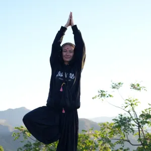 Female yoga practitioner performing tree pose (Vrksasana) with hands in prayer position outdoors against a mountain backdrop during 300 hour yoga teacher training in rishikesh.