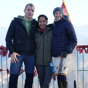 Three smiling yoga students wearing warm outdoor jackets and red tilak marks standing together during a mountain excursion for 50 hour yoga teacher training in rishi