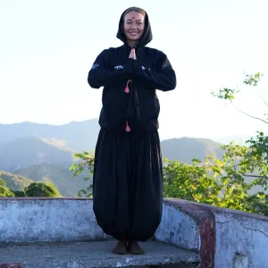 Yoga practitioner wearing a black hoodie standing in Namaste pose outdoors on a terrace with Himalayan mountains in the background during 50 hour yoga teacher training in rishikesh.